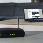 Wireless router with a tall antenna on a desk, green LED status lights lit, blurred laptop on a coffee table in the background.