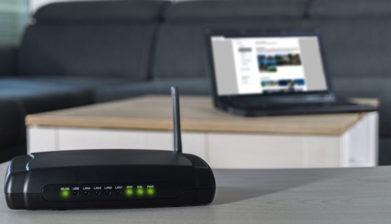 Wireless router with a tall antenna on a desk, green LED status lights lit, blurred laptop on a coffee table in the background.