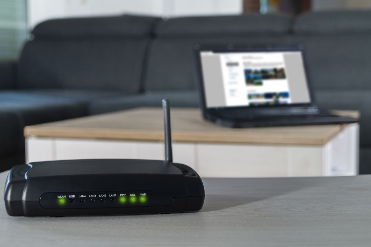 Wireless router with a tall antenna on a desk, green LED status lights lit, blurred laptop on a coffee table in the background.