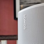 Close-up of a silver Sonos speaker grille with the brand name visible, set against a red wall and wooden railing.