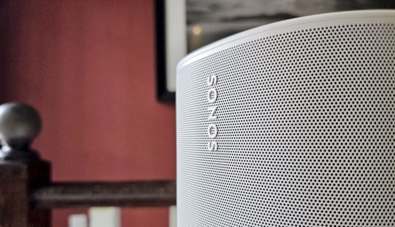 Close-up of a silver Sonos speaker grille with the brand name visible, set against a red wall and wooden railing.