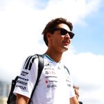 Young man wearing a white sponsor logo shirt and sunglasses stands outdoors against a bright blue sky and clouds, looking to the right with a backpack on. - or - A man in a white racing-style shirt with logos and a black backpack, wearing sunglasses, outdoors on a sunny day.