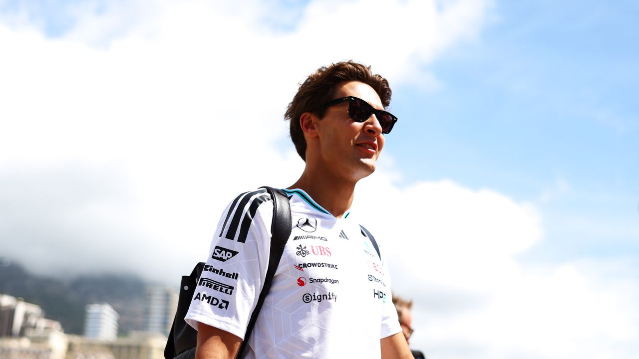 Young man wearing a white sponsor logo shirt and sunglasses stands outdoors against a bright blue sky and clouds, looking to the right with a backpack on. - or - A man in a white racing-style shirt with logos and a black backpack, wearing sunglasses, outdoors on a sunny day.