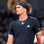 Male tennis player in a navy Adidas shirt and headband, looking to the right on a court with a blurred crowd in the background.