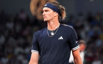 Male tennis player in a navy Adidas shirt and headband, looking to the right on a court with a blurred crowd in the background.