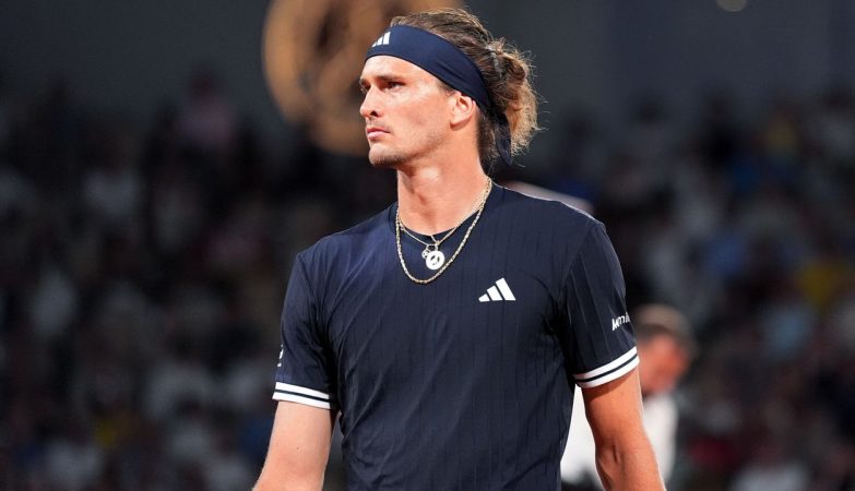 Male tennis player in a navy Adidas shirt and headband, looking to the right on a court with a blurred crowd in the background.
