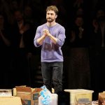 Man in a purple sweater stands on a stage among scattered cardboard boxes as the audience applauds in the darkened background.