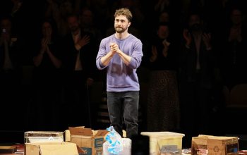 Man in a purple sweater stands on a stage among scattered cardboard boxes as the audience applauds in the darkened background.