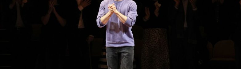 Man in a purple sweater stands on a stage among scattered cardboard boxes as the audience applauds in the darkened background.