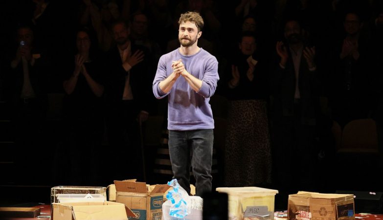 Man in a purple sweater stands on a stage among scattered cardboard boxes as the audience applauds in the darkened background.