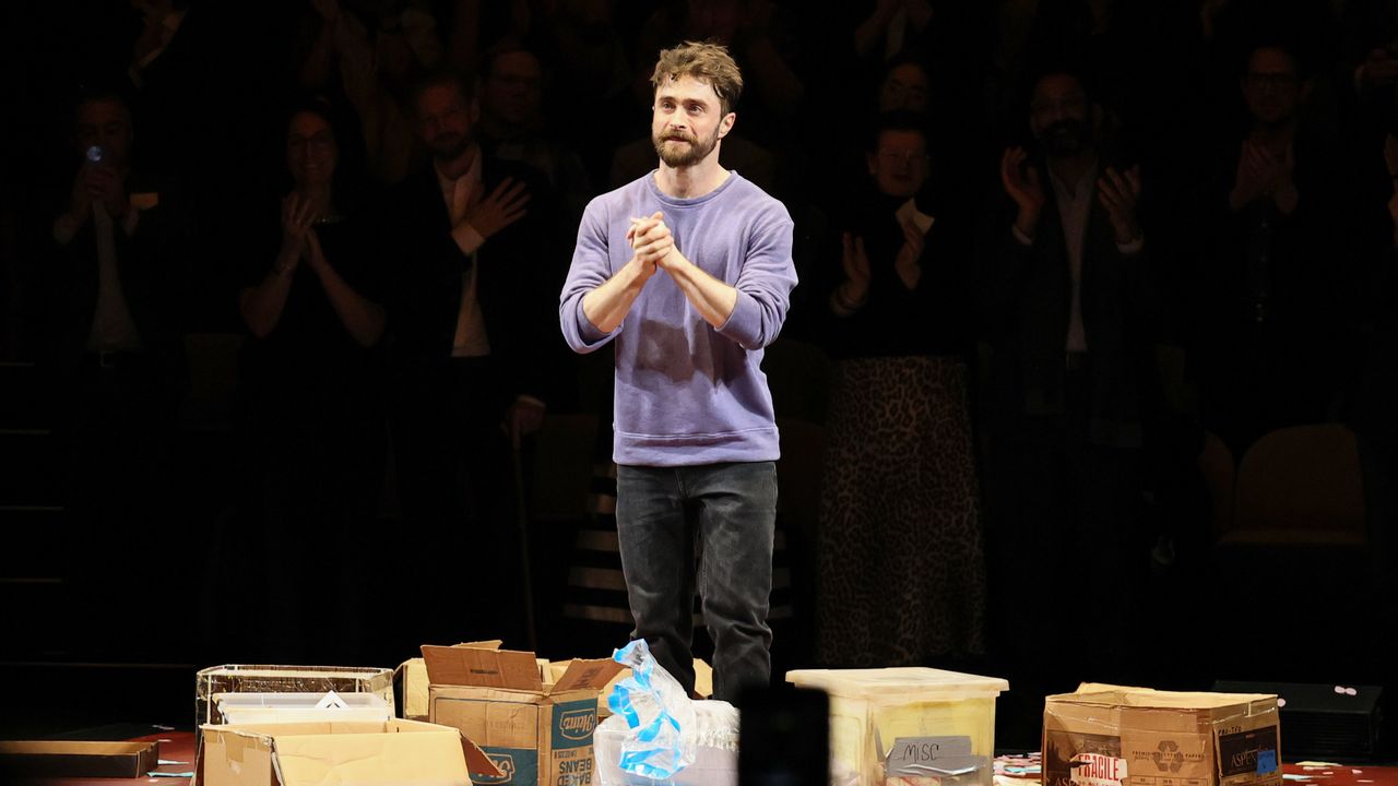 Man in a purple sweater stands on a stage among scattered cardboard boxes as the audience applauds in the darkened background.