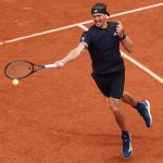 Male tennis player in a black outfit on a red clay court extends a forehand, racket reaching for a bright yellow ball just ahead of him on the court lines.