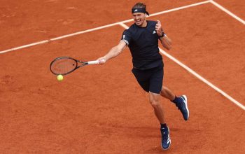 Male tennis player in a black outfit on a red clay court extends a forehand, racket reaching for a bright yellow ball just ahead of him on the court lines.