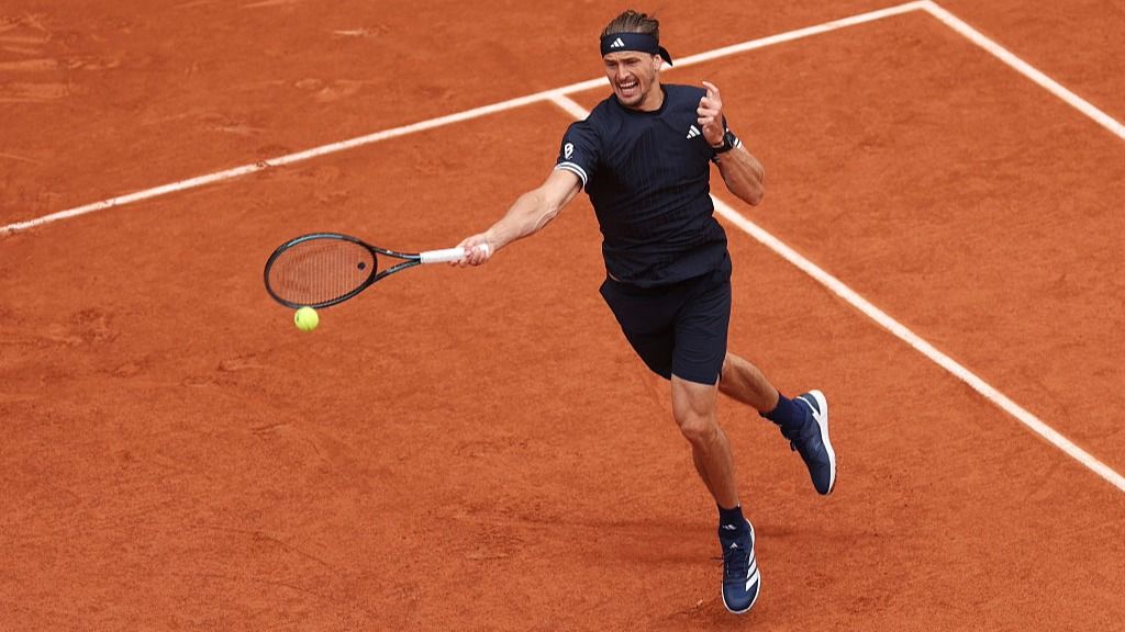 Male tennis player in a black outfit on a red clay court extends a forehand, racket reaching for a bright yellow ball just ahead of him on the court lines.