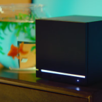 Black cube-shaped electronic device with a white glowing bar on the front, on a wooden surface beside a fishbowl.