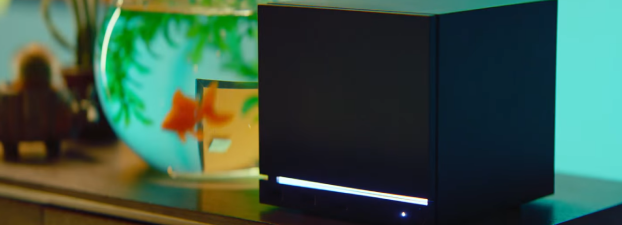 Black cube-shaped electronic device with a white glowing bar on the front, on a wooden surface beside a fishbowl.