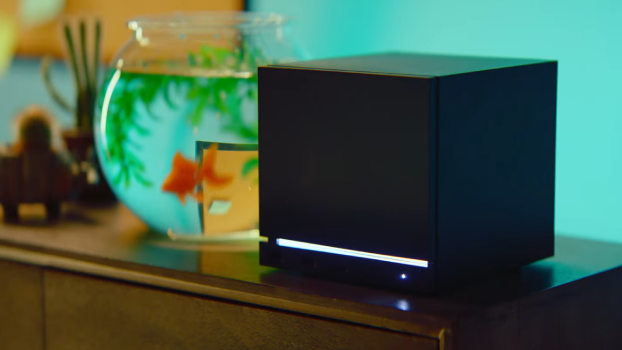 Black cube-shaped electronic device with a white glowing bar on the front, on a wooden surface beside a fishbowl.