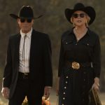 Couple in black western attire walking outdoors, both wearing cowboy hats and sunglasses.