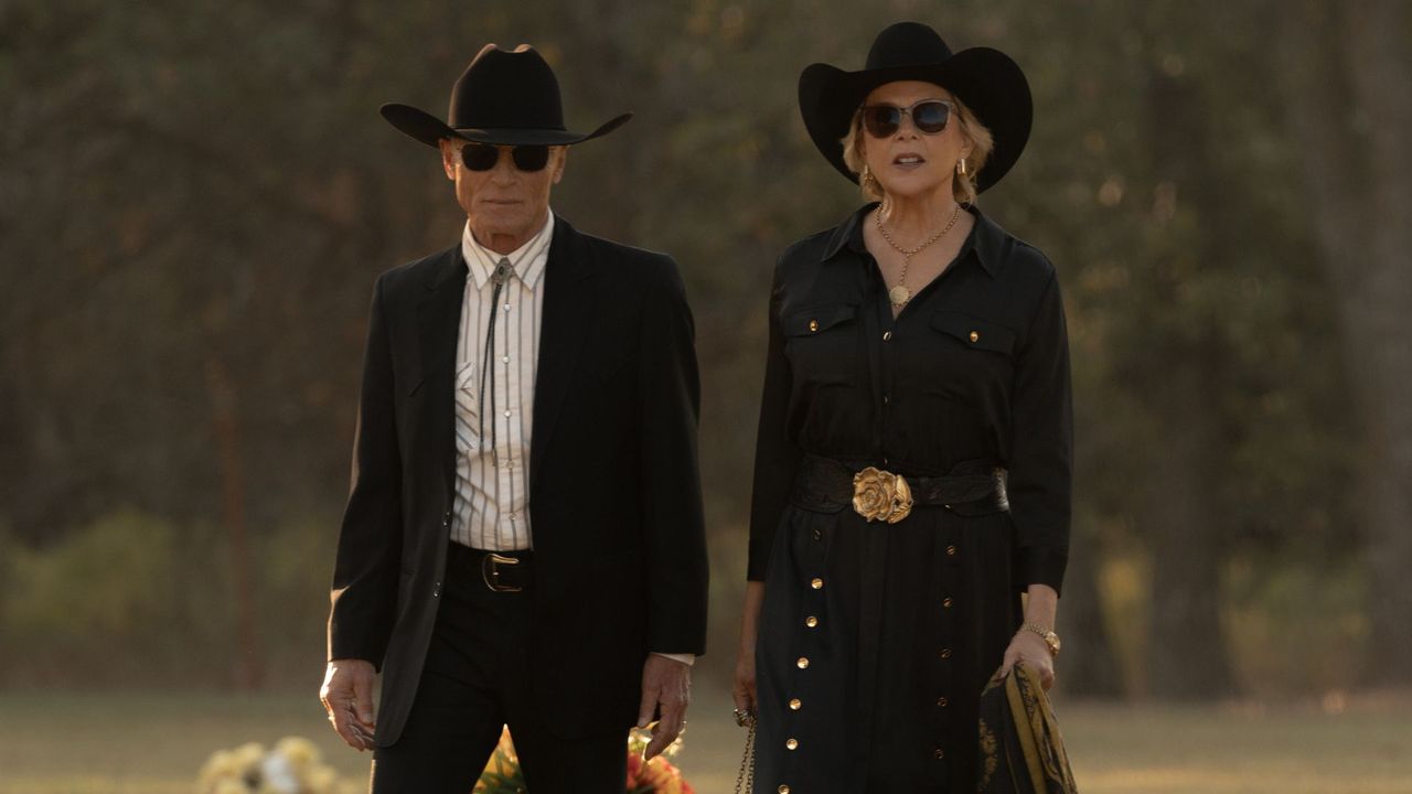 Couple in black western attire walking outdoors, both wearing cowboy hats and sunglasses.